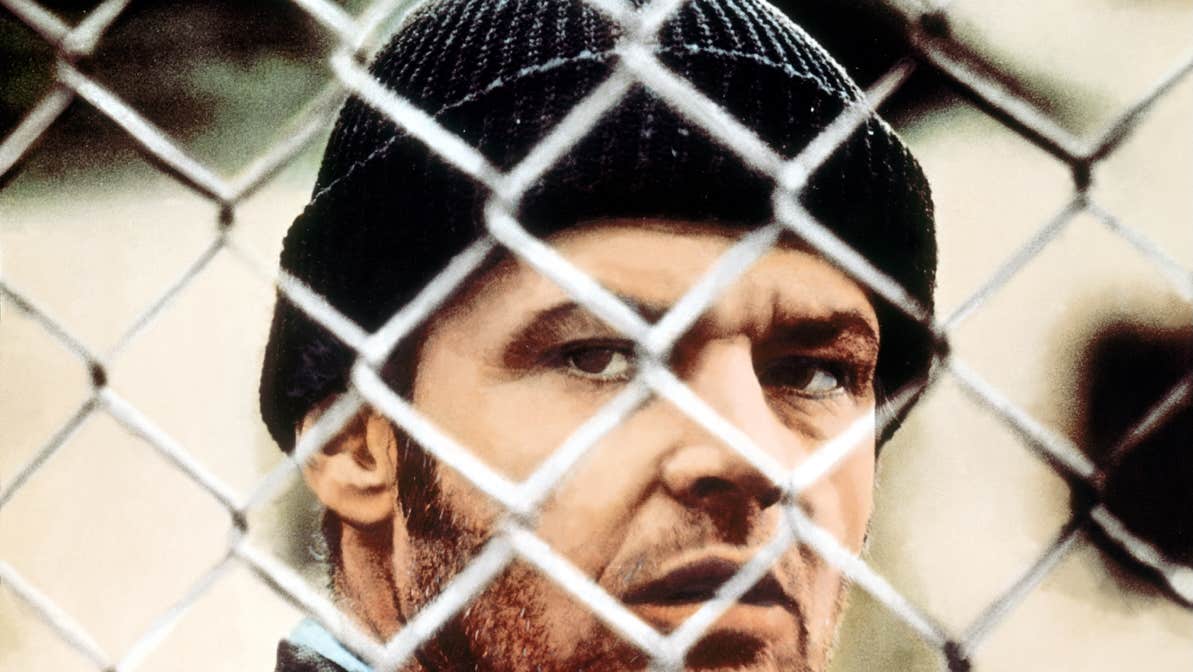 A gruff man is looking through a wire mesh fence.
