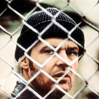 A gruff man is looking through a wire mesh fence.