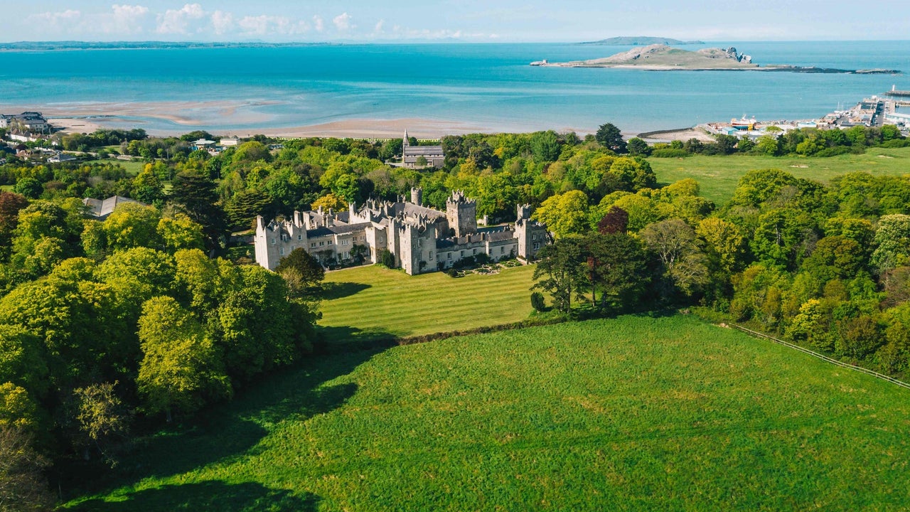 Aerial view of a castle surrounded by green fields and trees and a beach in the background