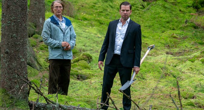A man in a crumpled raincoat and a man in a suit are in a forest.