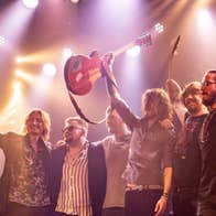 6 men in a line with arms around each other at the front of a stage, some holding up their arms and smiling.
