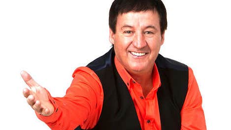 A smiling man in redshirt and black open waistcoat holding his right hand foreward.