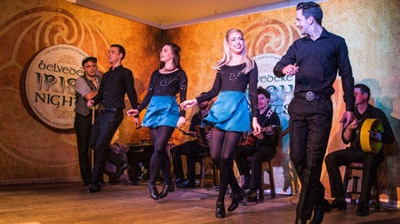 Four Irish dancers perform on a stage with traditional musicians seated behind them