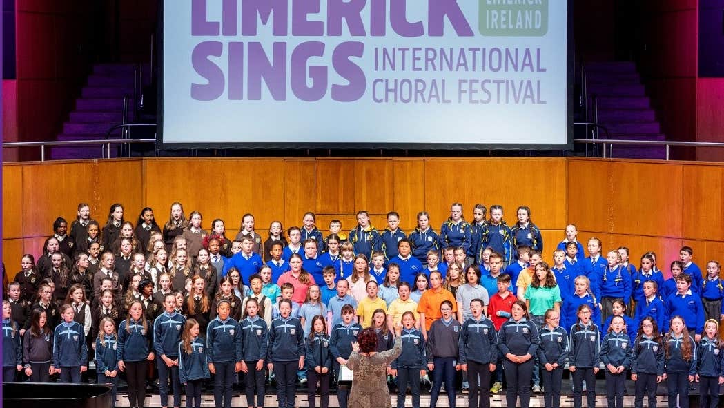 A distant view of a large stage with a big group of children singing on tiered steps with a conductor.
