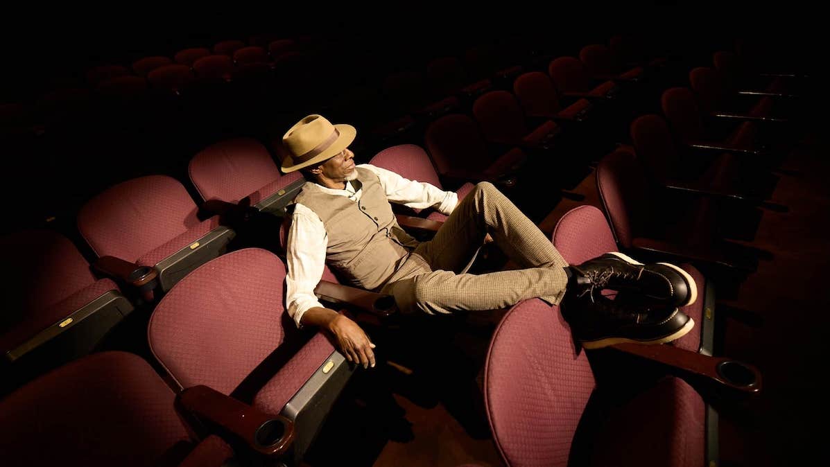 A man in waistcoat and shirt is seated in red cinema type seats with his feet up on the back on the seat in front, in darkness illuminated by a spot light.