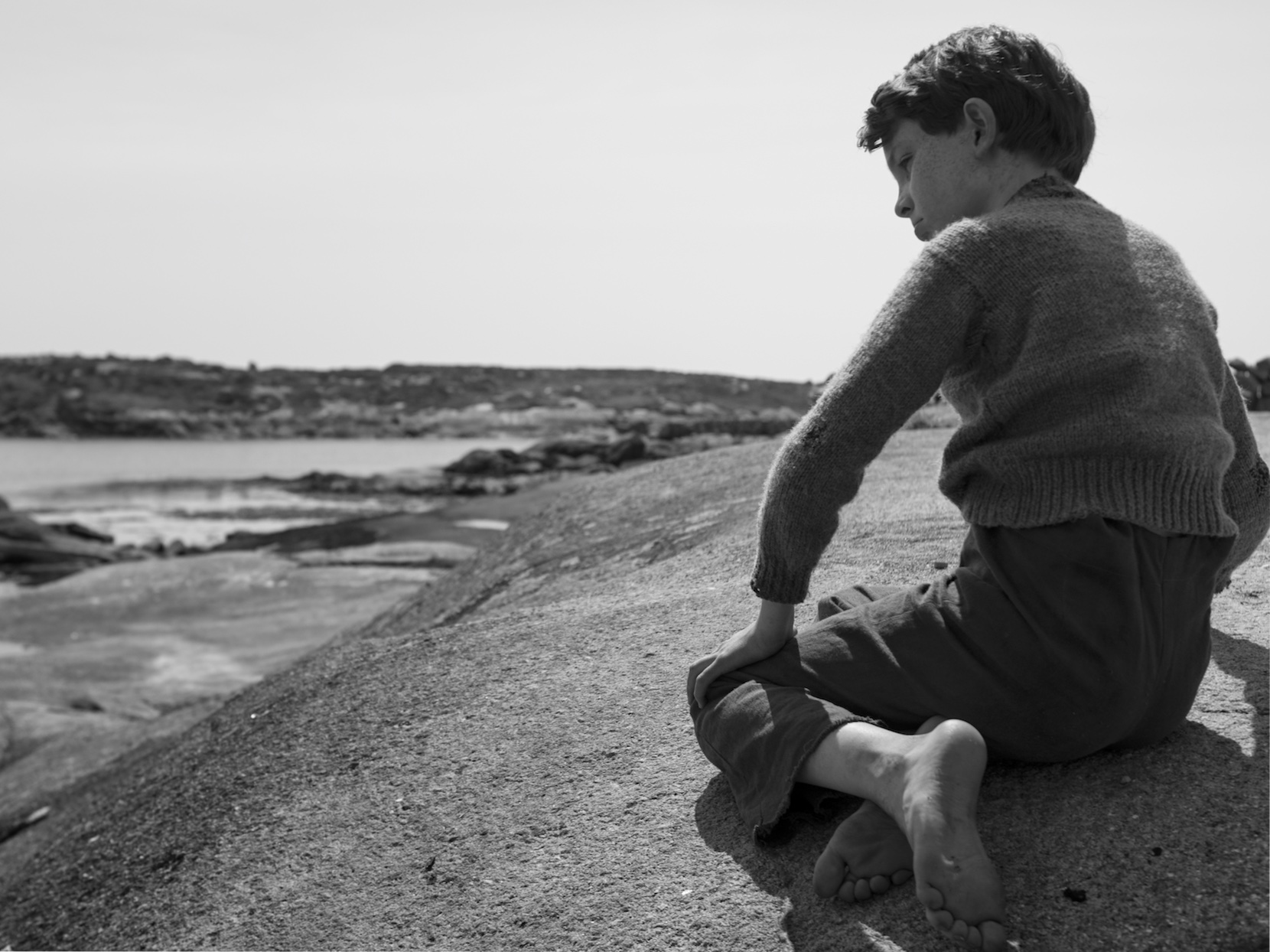 A boy is sitting on the ground near a river.