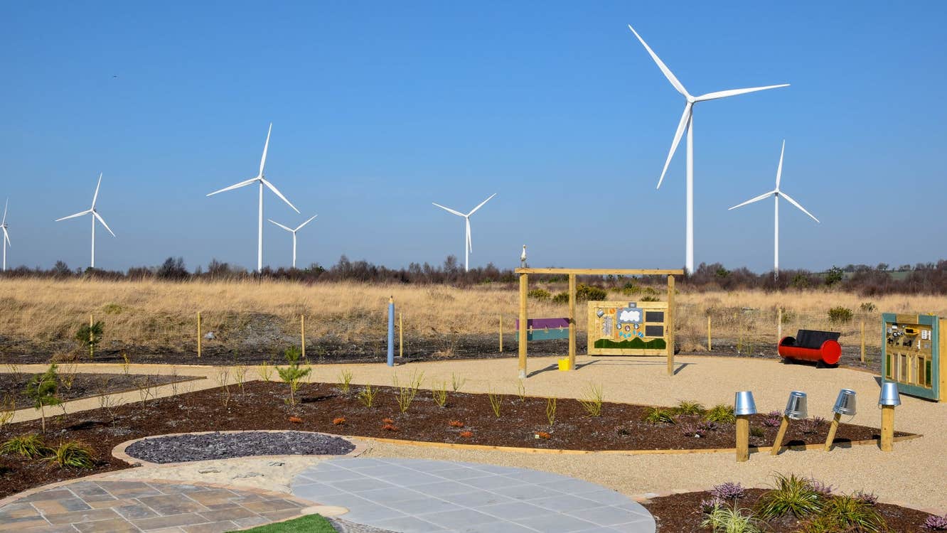 Paved area with plants grass a black and red bench and a wind farm in the background
