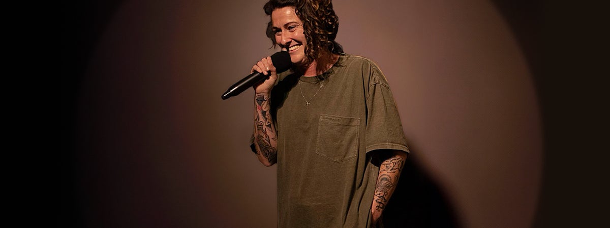 A smiling woman is holding a mic up to her mouth, against dark brown background.