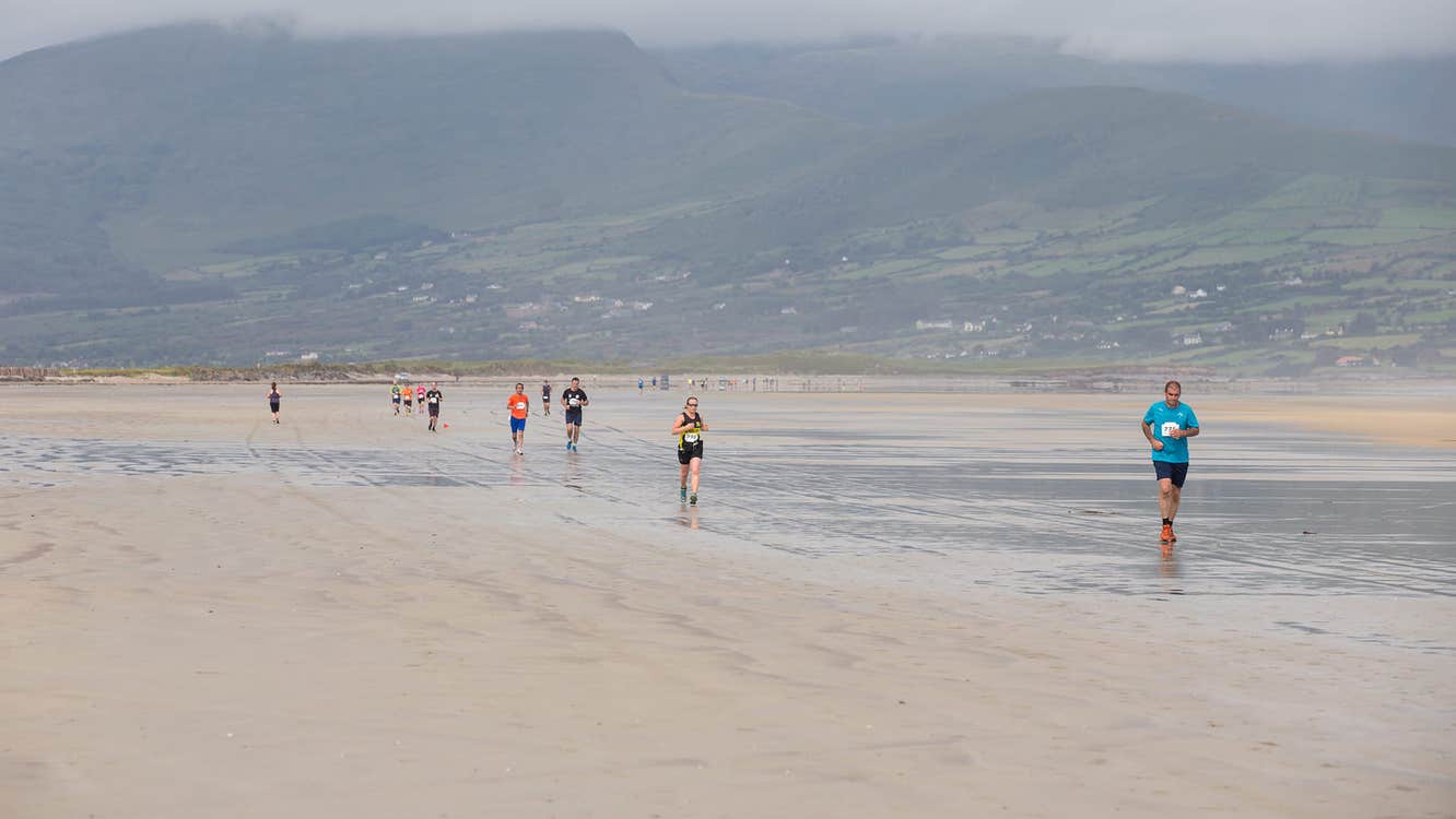 View of a large beach with people running along it, against background of hills