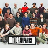 A large group of smiling men, some seated, the rest standing, against plain white background.