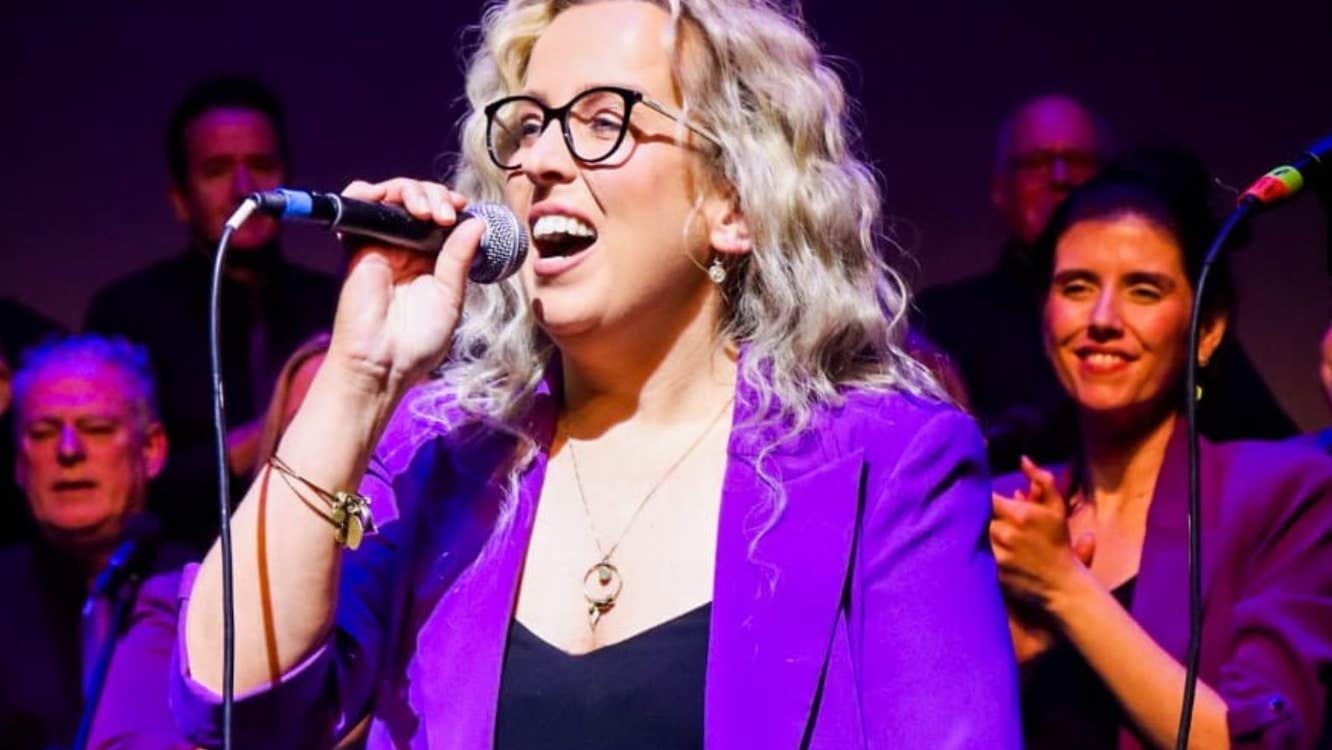 A woman in purple jacket singing with a hand held mic, with audience behind.