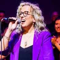 A woman in purple jacket singing with a hand held mic, with audience behind.