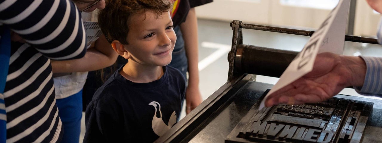 A young boy with his parents behind him looks over a hand rolling proofing press as the guide demonstrates how it works