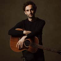 A man in black clothes sitting on a stool, resting his arms on a brown guitar.