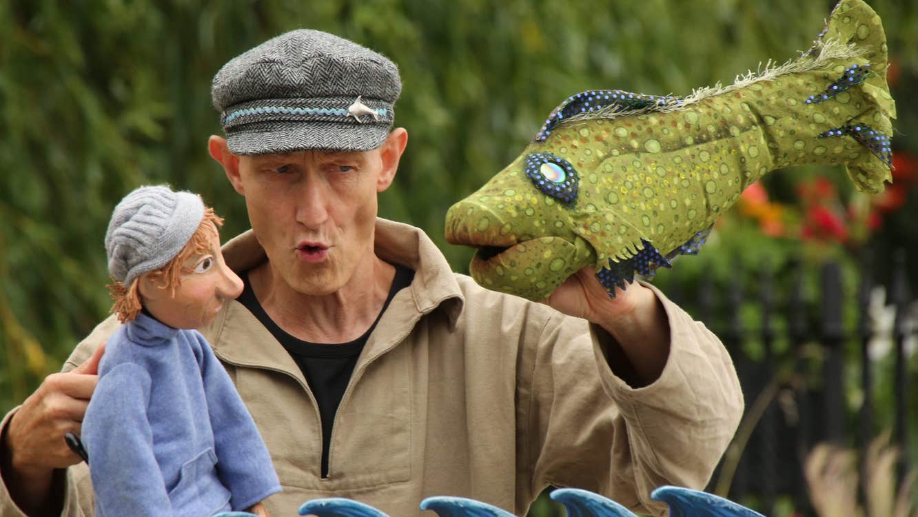 A man holding a puppet of a person in one hand looking at a large green fish puppet in the opposite hand.