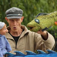 A man holding a puppet of a person in one hand looking at a large green fish puppet in the opposite hand.