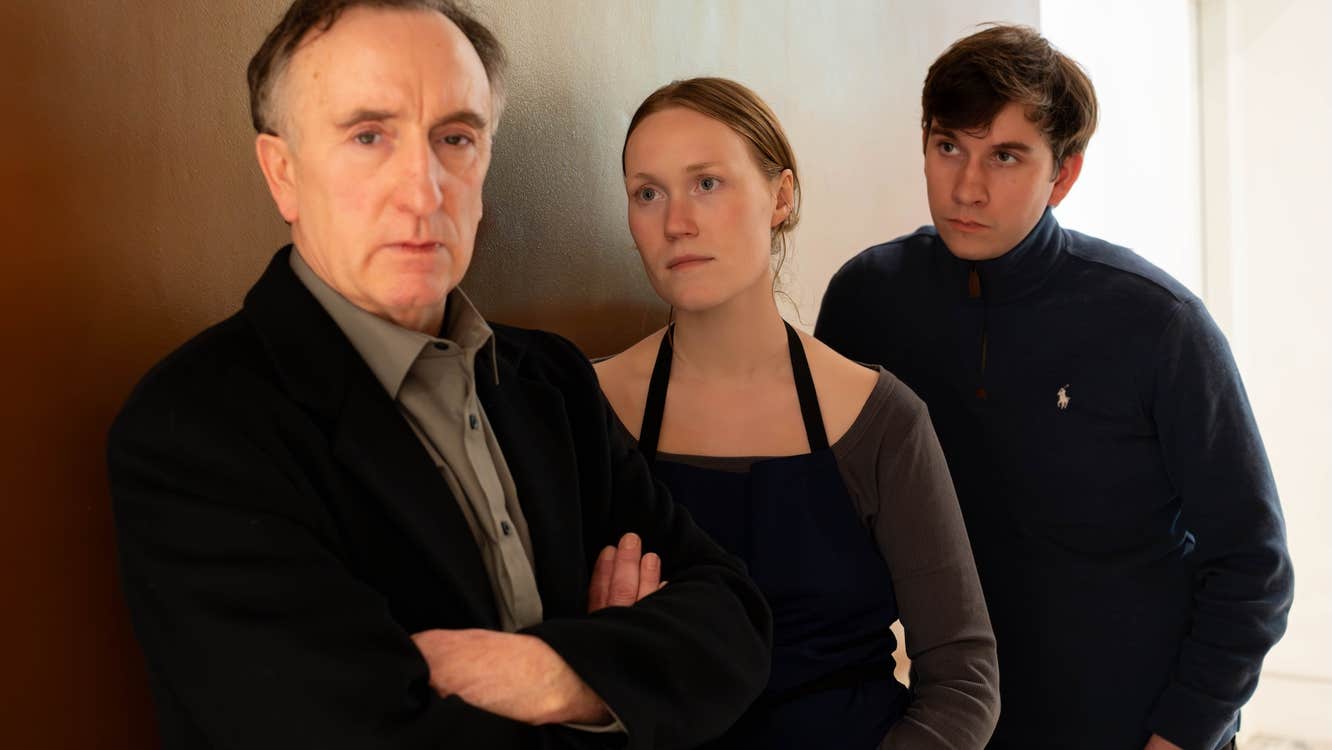 All leaning against an internal wall, 2 people are looking at a man with crossed arms, not looking at them.