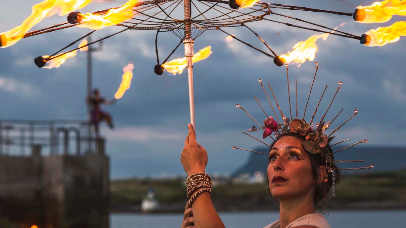 A frame of an umbrella with flames at the end of each of the spokes is being held upright by a person wearing a tall, spiked headdress against dark, cloudy background of a quay and distant mountain.