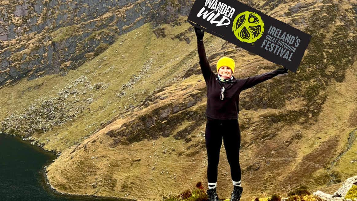 A person dressed in dark outdoor clothing is standing on a rock holding up a large black banner with event logo and name on.