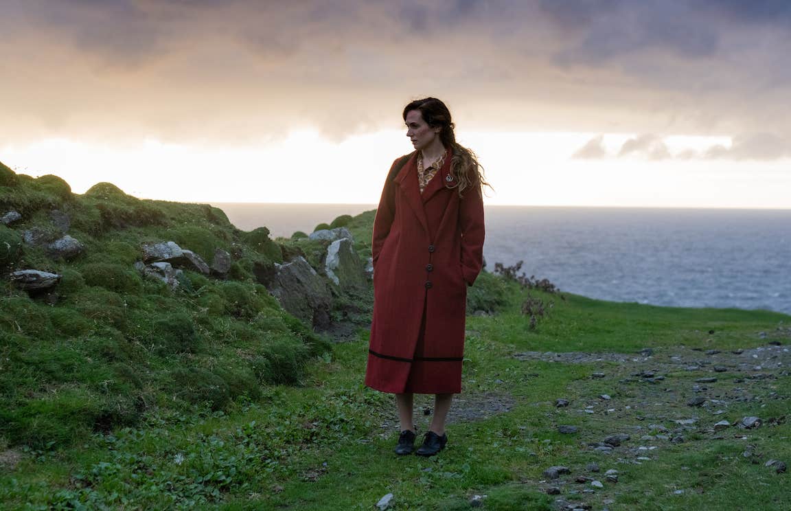 A scene from the movie, 'The Banshees of Inisherin' featuring actress Kerry Condon in a red coat, standing in a field with the Atlantic Ocean in the background.
