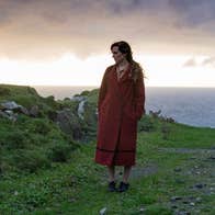 A scene from the movie, 'The Banshees of Inisherin' featuring actress Kerry Condon in a red coat, standing in a field with the Atlantic Ocean in the background.