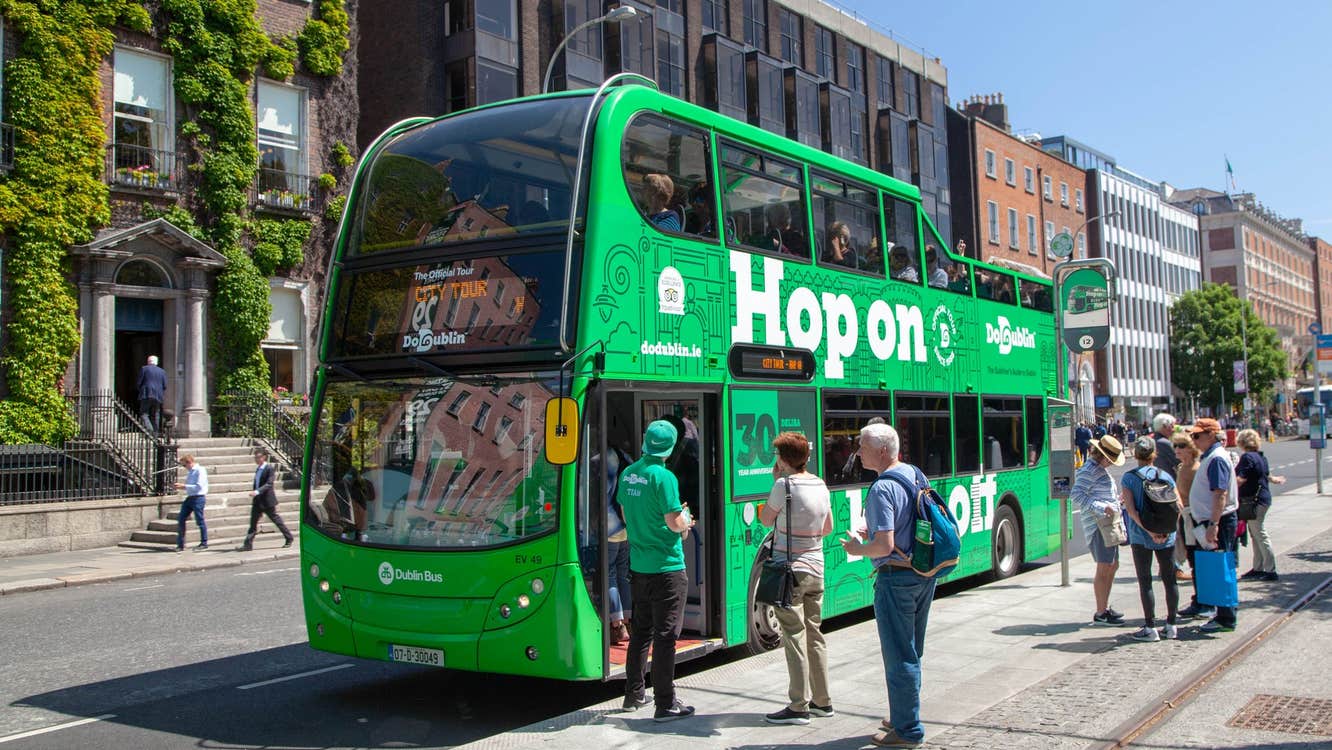 A large green double decker bus with people queueing to get on it
