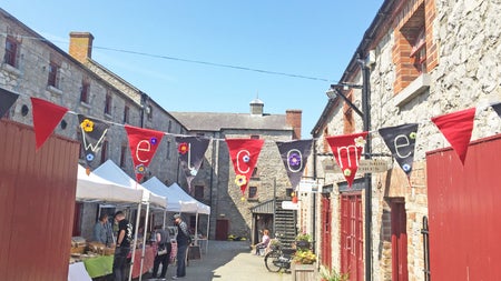 Skerries Mills Farmers Market stalls in a lane next to old stone buildings and a welcome sign