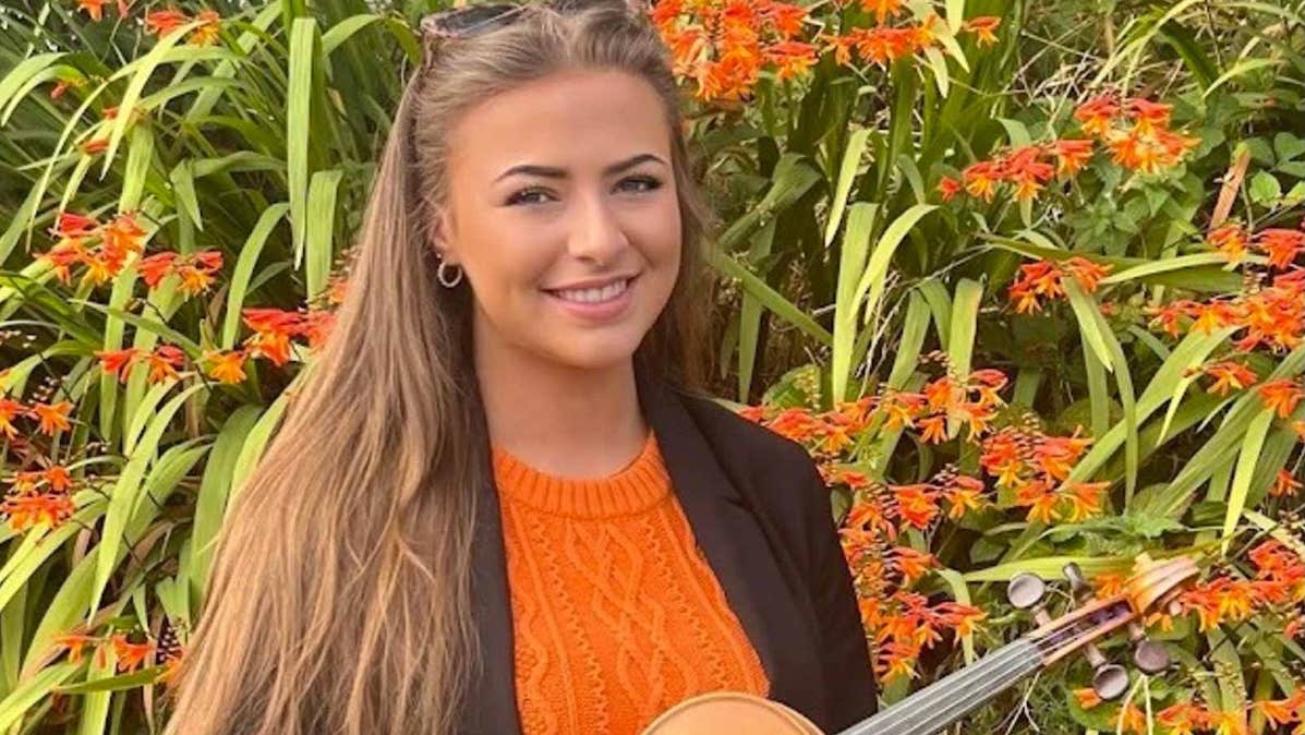 A smiling young woman holding a fiddle with large flowering plant in background.
