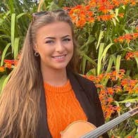 A smiling young woman holding a fiddle with large flowering plant in background.
