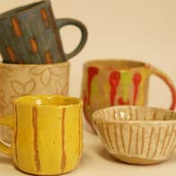 A group of 5 small, rustic handmade mugs in shades of yellow, beige, red and dark green.
