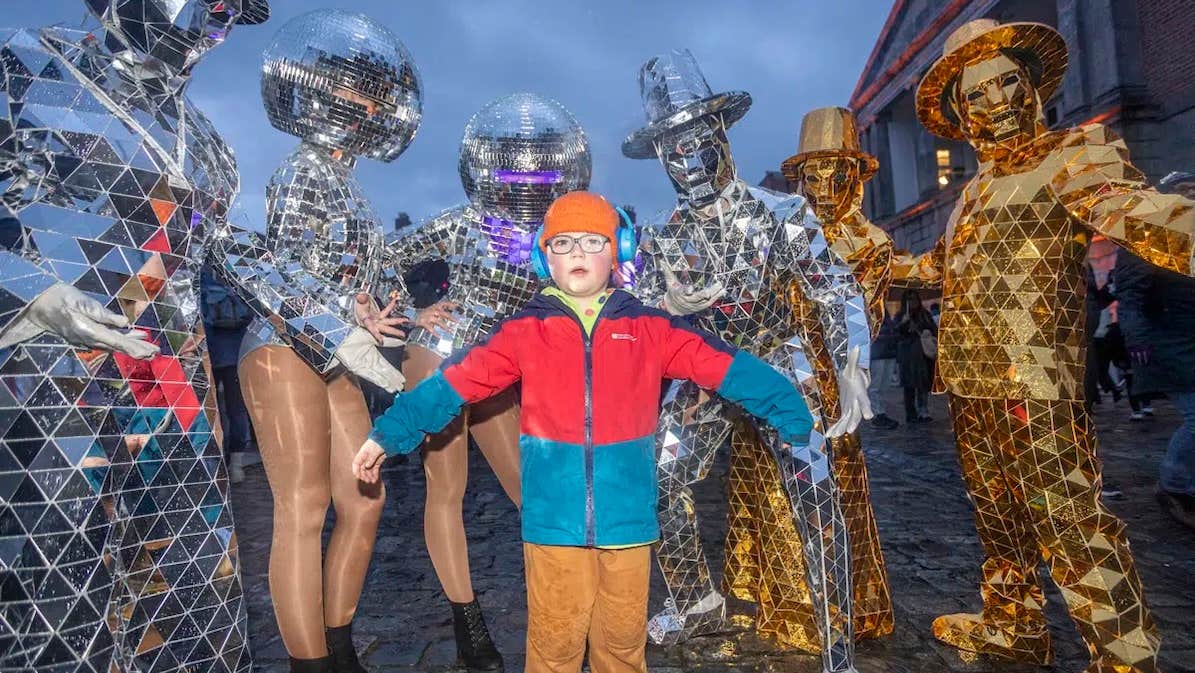 A child outdoors in coat and hat standing with group of 6 people in silver or gold mosaic covered costumes with faces hidden by headwear.