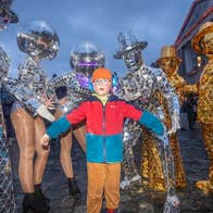 A child outdoors in coat and hat standing with group of 6 people in silver or gold mosaic covered costumes with faces hidden by headwear.