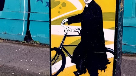 Street art of Michael Collins wearing a long black coat and hat with his bicycle on a yellow background