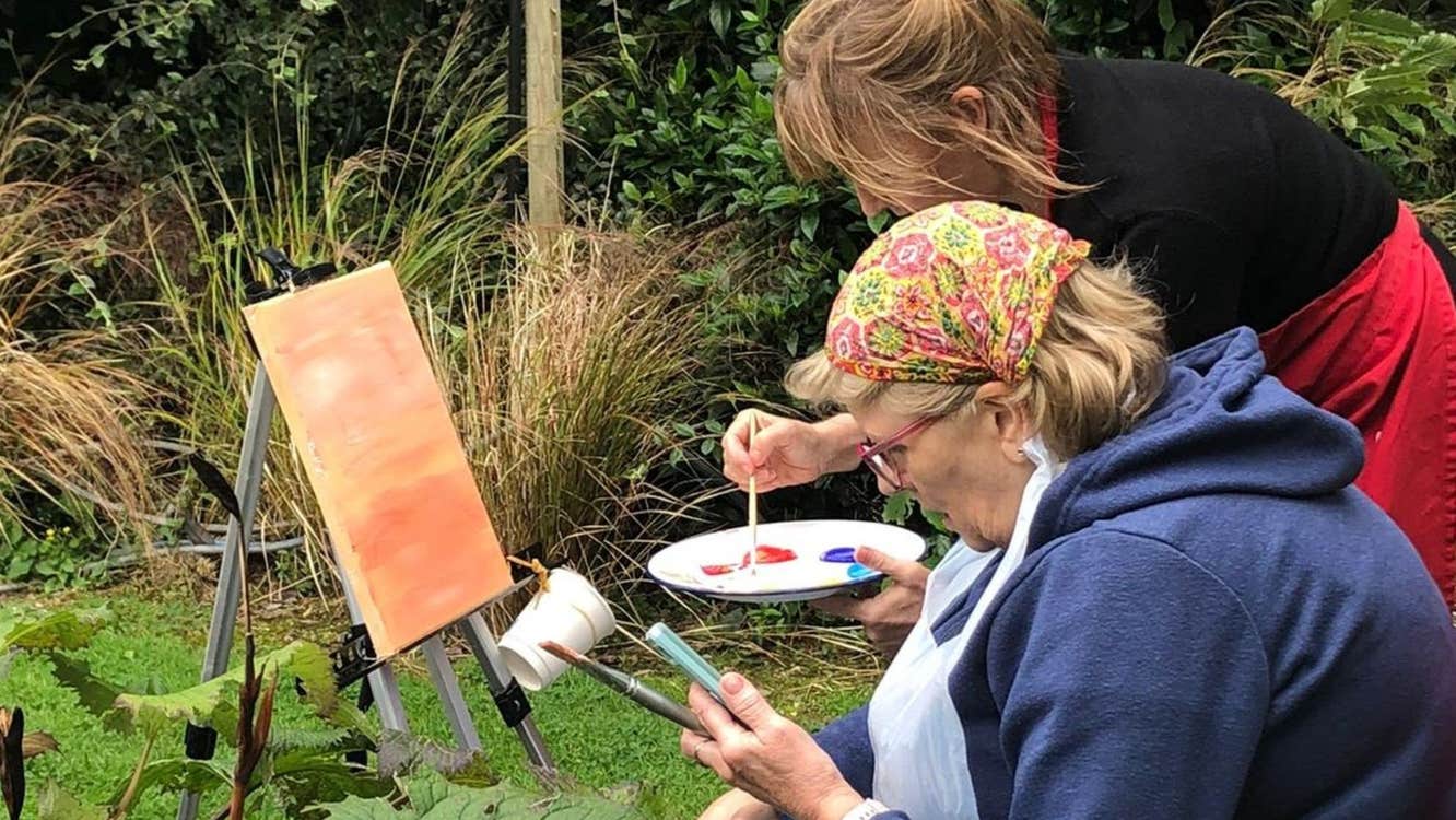 An artist sitting outdoors at an easel while another artist mixes paint on a palette