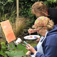 An artist sitting outdoors at an easel while another artist mixes paint on a palette