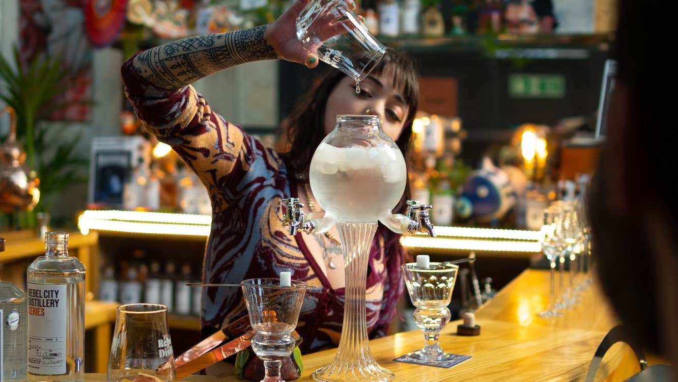 A woman is behind a bar pouring liquid into a very tall slim glass with a large, bulb shaped top.