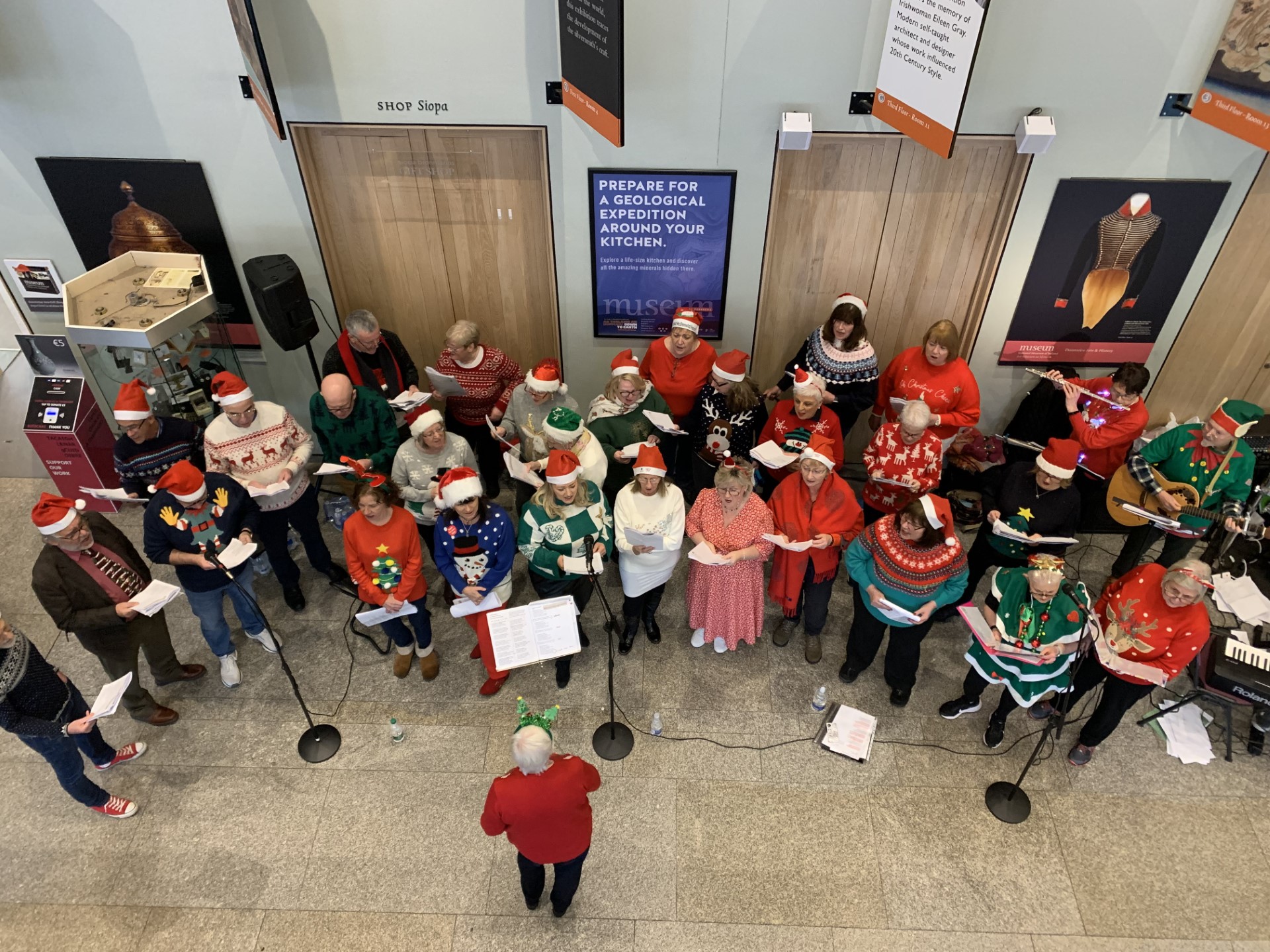 St. Michael's Reunion Choir Performance, a small choir standing in a semi circle with a conductor at the front facing them, seen from above looking down, some wearing christmas jumpers or santa hats.
