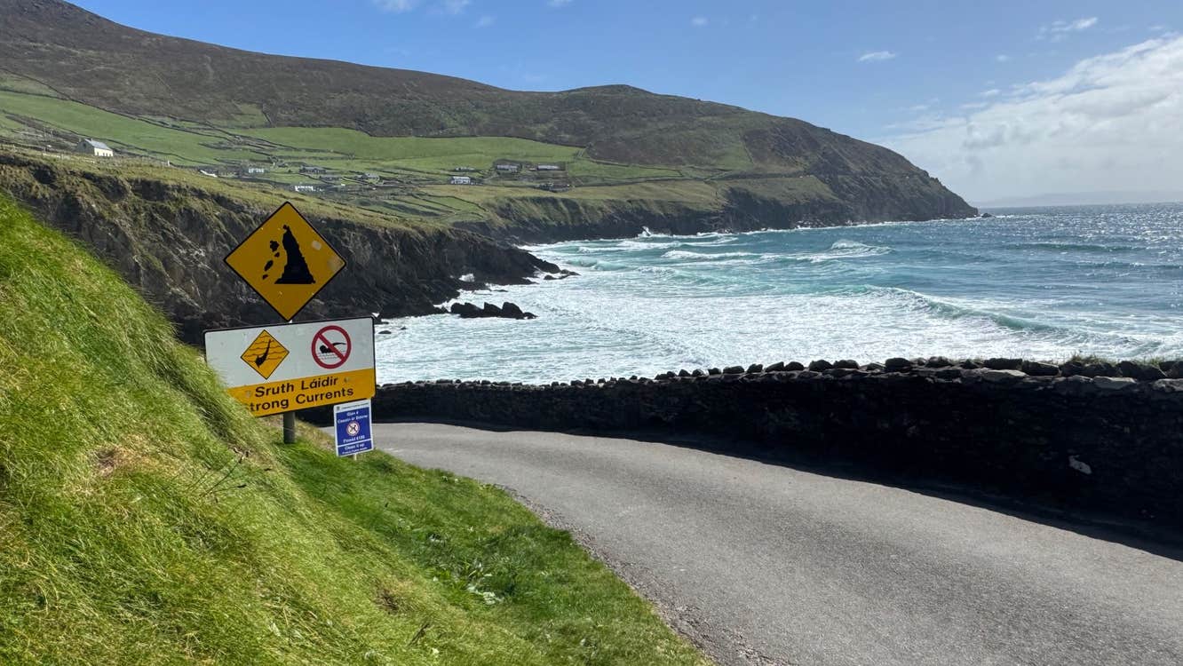 Coastal road overlooking waves crashing against cliffs under a bright sky with warning signs posted along the roadside