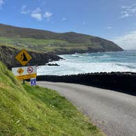 Coastal road overlooking waves crashing against cliffs under a bright sky with warning signs posted along the roadside