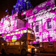 At night a vintage double decker bus is parking outside a large out building the front of which is covered in pink lights with white snowflakes.