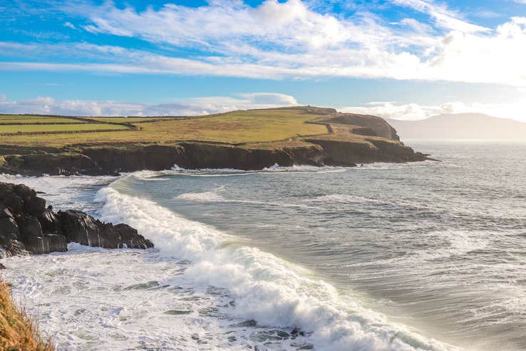 Scenic photo of Dingle Bay county Kerry, featuring a headland and crashing waves, with blue skies.