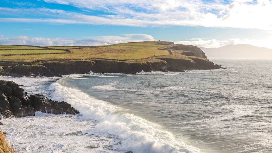 Scenic photo of Dingle Bay county Kerry, featuring a headland and crashing waves, with blue skies.