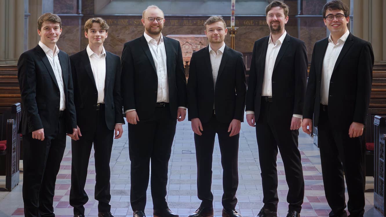 6 smiling men in dark suits and white shirts a in a line in a wide church aisle