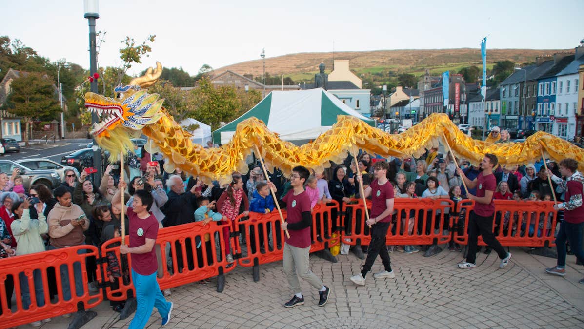 Young men walking in a line holding up poles attached to a yellow and red dragon puppet in front of a crowd who are behind red barriers, outside in a town square.