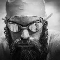 Close up headshot of man with a beard wearing swimming goggles and hat, covered in water droplets.
