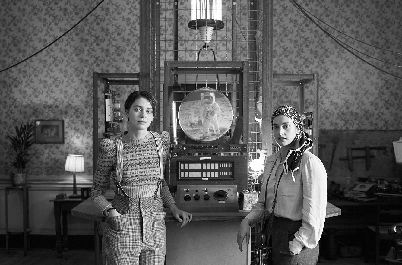 Black and white image of 2 women standing in front of an old fashioned style machine with a round screen showing the moon landing.