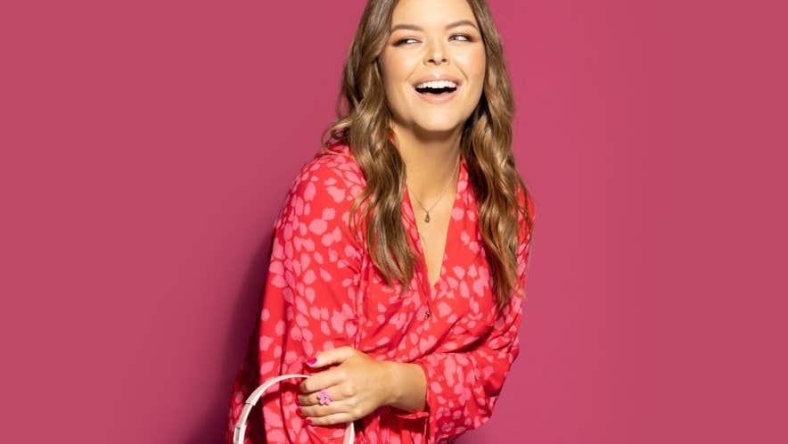 A laughing woman in a red dress holding a pair of overear headphones against plain dark pink background.