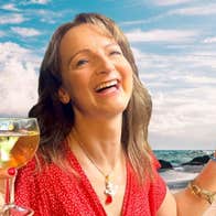 A laughing woman holding a glass of wine with beach in the background.