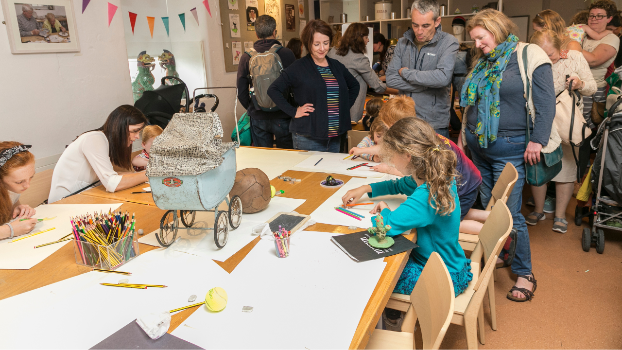 Children are seated at a table drawing on large sheet of white paper with adults looking on.