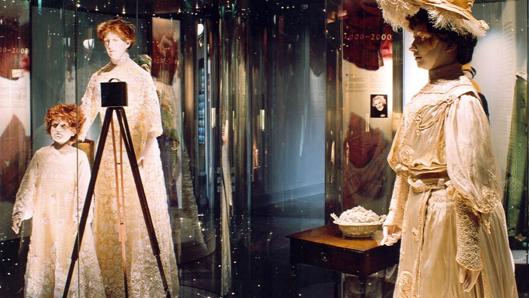 Display cabinets containing life size models wearing old clothing in beige colours in dimly lit room.